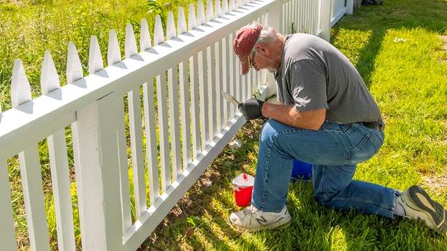A man bends at the knee as he paints a white picket fence. A small white barn is nearby.