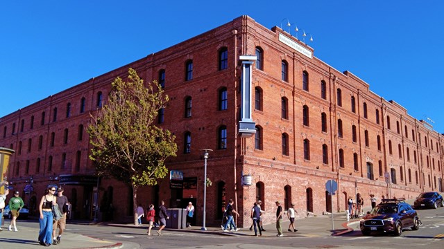 A historical brick warehouse stands on the corner of a busy street on a sunny day.