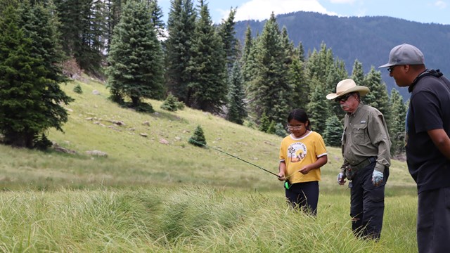 Two adult mentors show a young girl how to flyfish on a mountain stream.